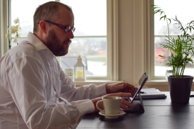 50 yaşında, beyaz tişörtlü, serbest sakallı bir adam bir masada oturur ve bir dizüstü bilgisayara not alır. Konsept: Evde dizüstü bilgisayarın arkasında serbest çalışan, karantinada çalışan.