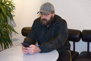 Gözlüklü sakallı bir adam halka açık bir masada oturuyor ve elinde bir akıllı telefon tutuyor..
