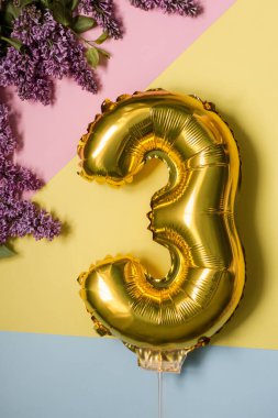 Üç yıllık doğum günü. Eflatun çiçekli renkli arka planda 3 numaralı altın folyo balon çiçek açıyor. Yıldönümü, kutlama konsepti. Düz yatış, üst görünüm