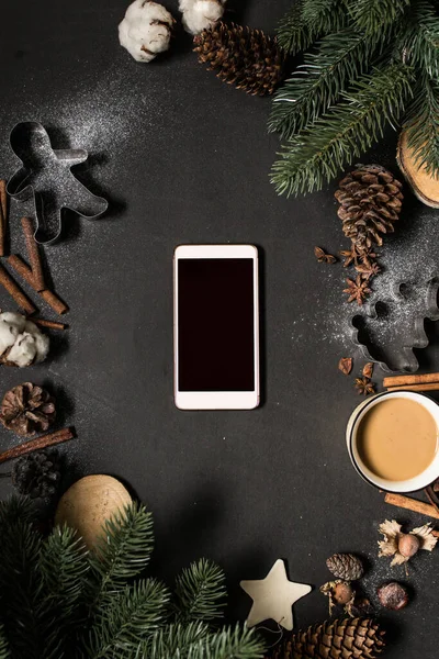 Christmas holidays spirit concept: smartphone with pine twigs, cones, cinnamon sticks, anise stars, cup of hot drink, biscuit forms and snow frame on black. Creative flat lay, blank display for copy.