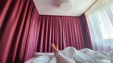 Kadın bacakları, temiz beyaz çarşaflarla yatakta uzanıyor. Sabah yatak odasında rahatla. Arka planda Burgundy perdeleri var. Sabahları rahat bir yatak geniş açılı.