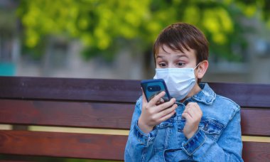 Karantina covid-2019 açık havada. Tıbbi maskeli bir çocuk elinde akıllı bir telefonla parktaki bankta oturur ve ekrana bakar. Şaşırdı, mutlu, heyecanlı ve neşeli..