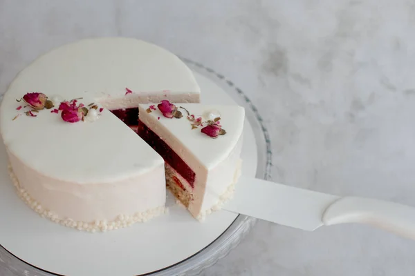 ミルククリームムースとラズベリーの入った白いケーキのピースは ラウンド透明ガラススタンドのカットケーキの横に白い肩のブレードに充填します 白い滑らかな釉薬とケーキ乾燥バラの芽 — ストック写真