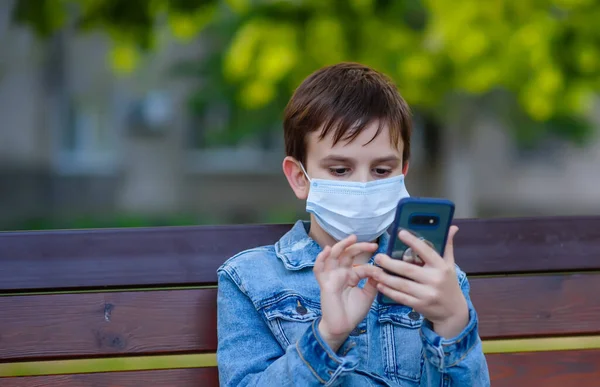 A boy in a medical mask and jeans clothes sits on a park bench with a smartphone in his hand and touching the screen. He is playing game or chatting
