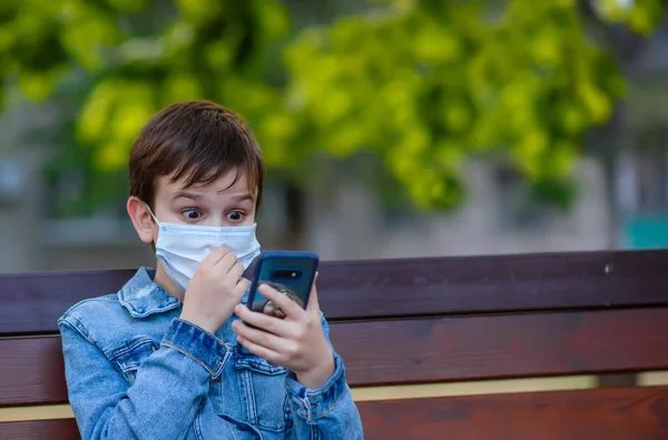 Happy and surprised boy 9-10 years old  in a medical protective mask sits on a  bench in the park and looking at the screen of smartphone.