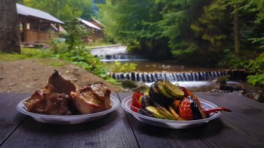 Izgarada pişmiş sebzeler ve tek kullanımlık yemeklerde ızgara et bir dağ nehrindeki şelalenin zeminine ve ormandaki büyük köknar ağaçlarına karşı. Güvenli Kafe. Sosyal uzaklık. Sağlıklı beslenme. Dağlara yolculuk.