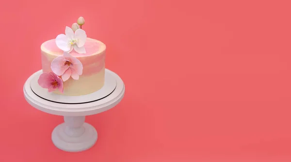 Pink marble cake stands on a round white stand on a pink-peach background. Cake decorated with sugar flowers orchids. Beautiful dessert decorated with flowers.