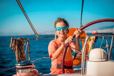 A handsome boy of 9-10 years old at the helm of a yacht in bright orange clothes, blue glasses against a blue sky. Sea vacation concept, sailing regatta. Traveling with children. clipart