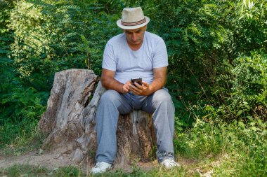 Orta Çağ Adamı Büyük Yaşlı kütüğün üzerinde oturuyor ve Doğada Akıllı Telefonla Oynuyor