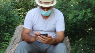 Yüzünde Tıbbi Maskeli Orta Çağ Adamı Doğada Oturuyor ve Akıllı Telefonuyla Oynuyor