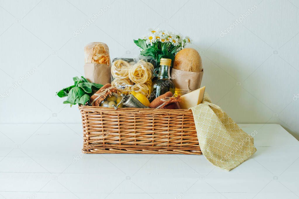 Italian food basket with ciabatta bread, tagliatelle, basil, olive oil, marinated olives in a mason jar, lemon, cheese, pepper mill, napkin, and a bouqet of chamomiles.