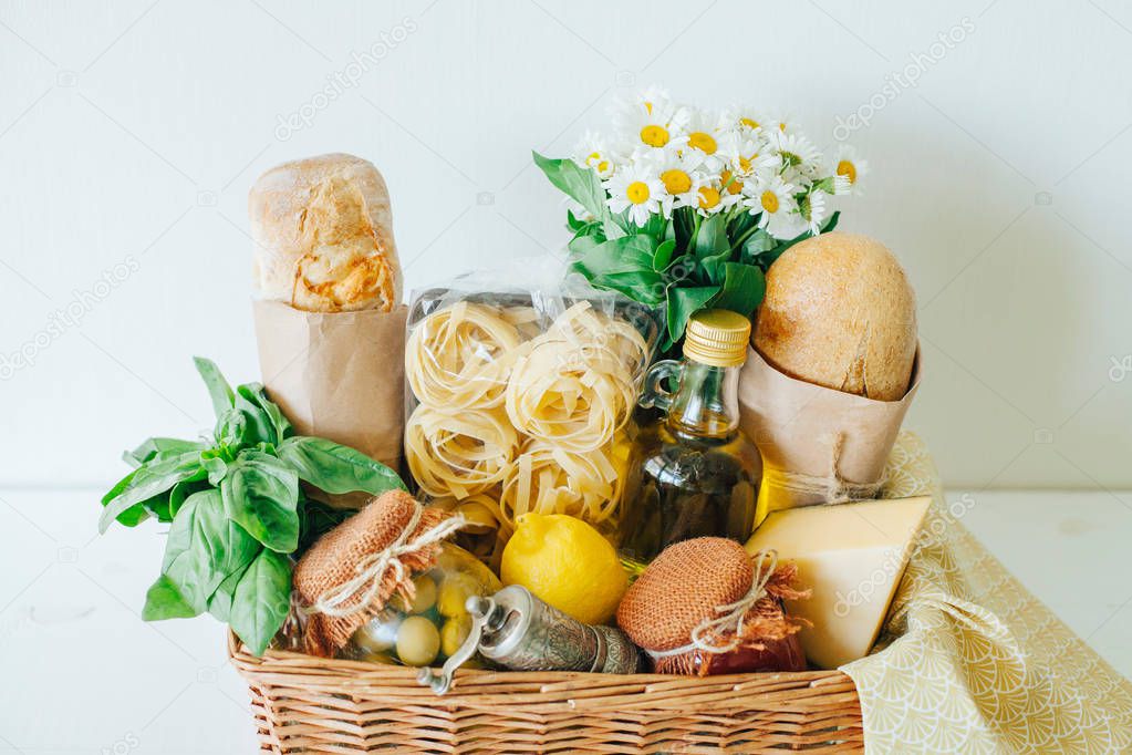 Italian food basket with ciabatta bread, tagliatelle, basil, olive oil, tomato sauce and marinated olives in a mason jar, lemon, cheese, pepper mill, napkin, and a bouqet of chamomiles.