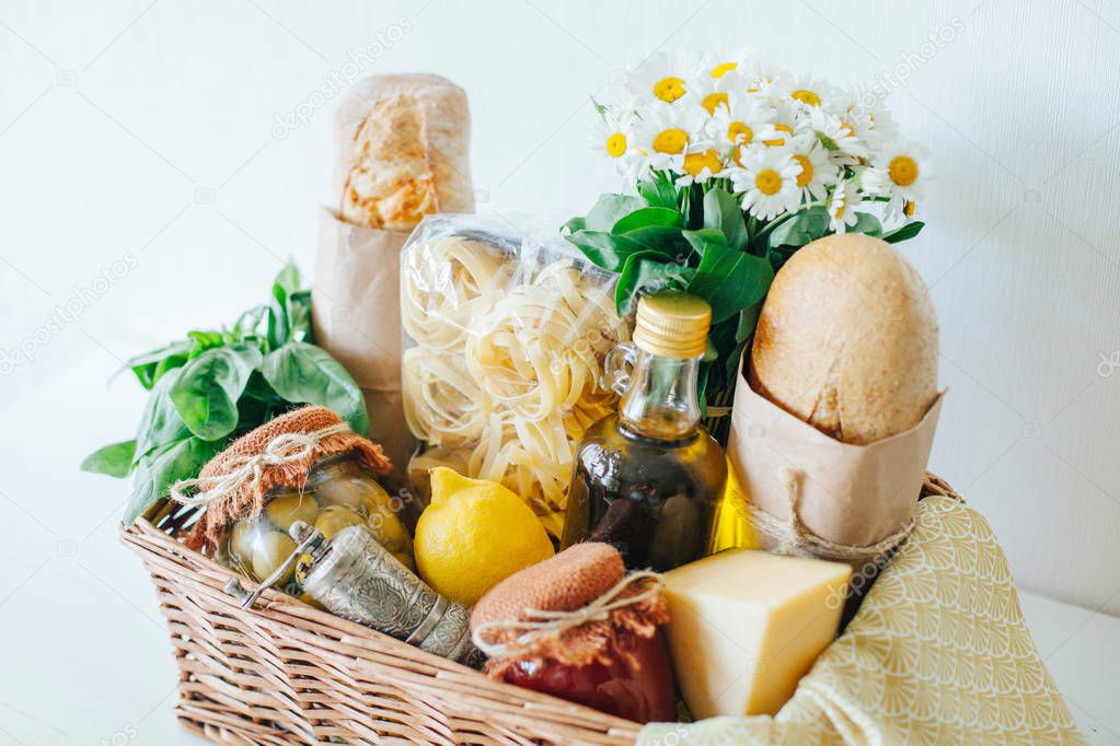 Italian food basket with ciabatta bread, tagliatelle, basil, olive oil, pickled olives and tomato sauce in a mason jar, lemon, cheese, pepper mill, napkin, and a bouqet of chamomiles.