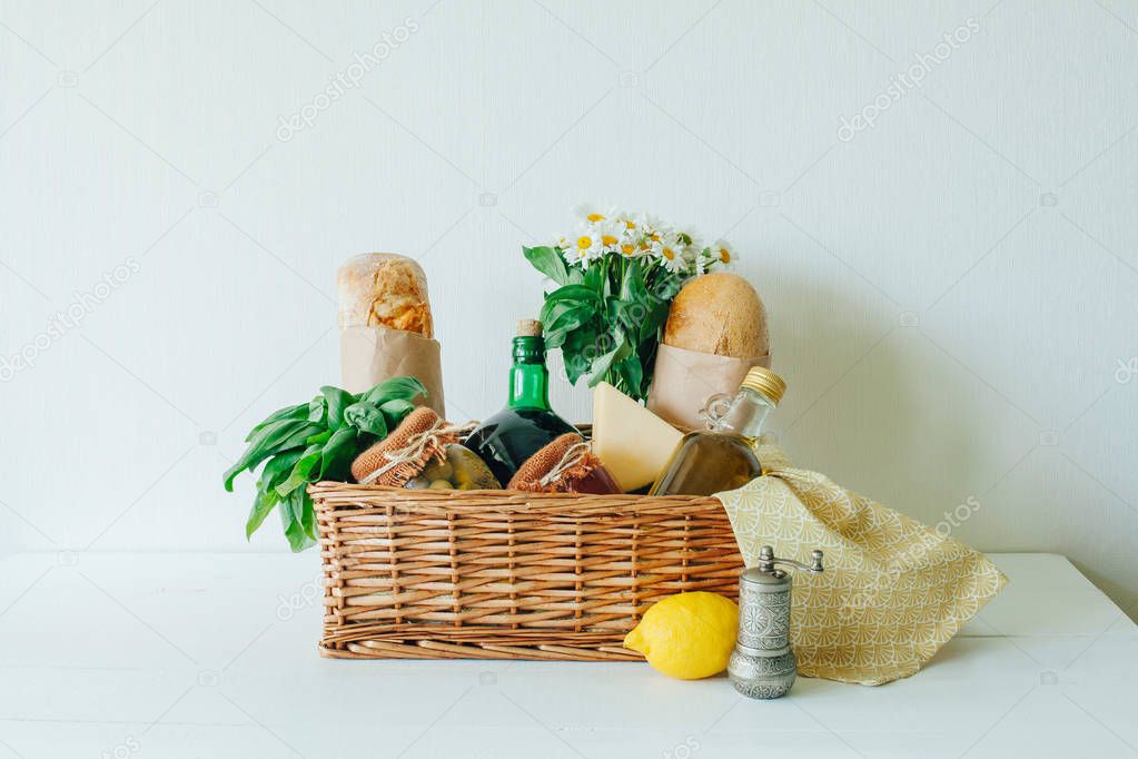 Italian food basket with ciabatta bread, tagliatelle, basil, olive oil, marinated olives in a mason jar, bottle of wine, lemon, cheese, pepper mill, napkin, and a bouqet of chamomiles.