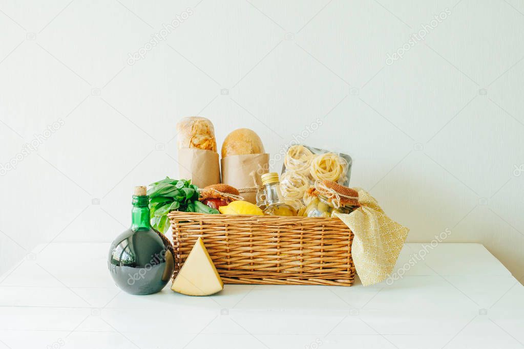 Italian food basket with ciabatta bread, tagliatelle, basil, olive oil, marinated olives in a mason jar, lemon, napkin, cheese and a bottle of wine.