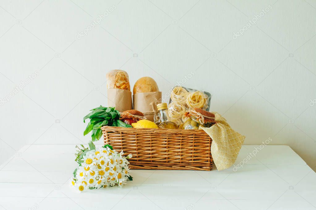 Bouqet of chamomiles and Italian food basket with bread, tagliatelle pasta, basil, olive oil, pickled olives, lemon, napkin, and a jar of tomato sauce.