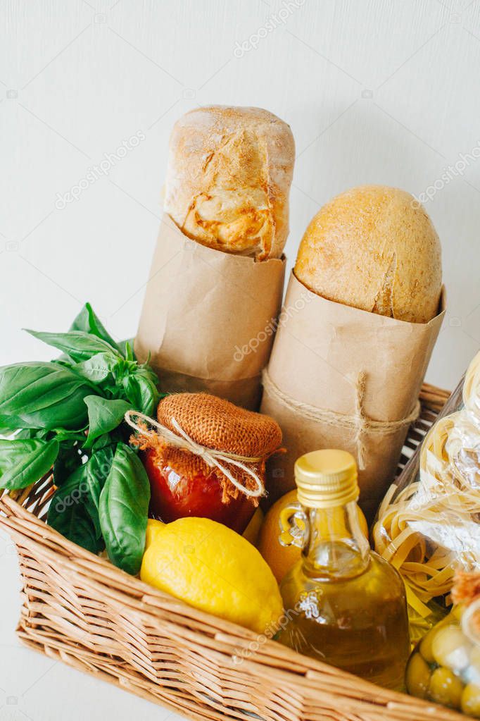 Italian food basket with ciabatta bread, tagliatelle, basil, olive oil, pickled olives, lemons, and a jar of tomato sauce.
