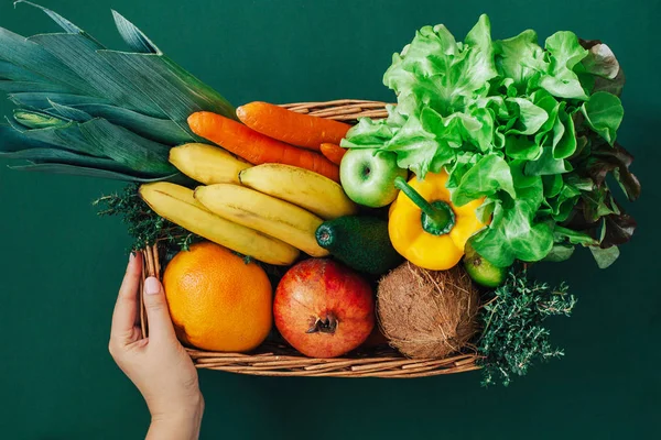 Wicker basket with fresh vegetables and fruits on green background