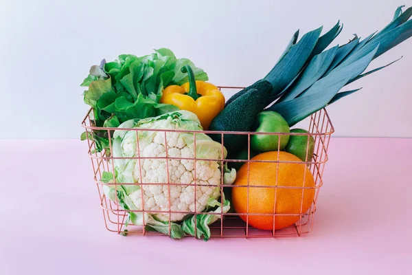 Basket with green fruits and vegetables on pastel pink background