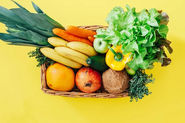 Wicker basket with fresh vegetables and fruits on yellow background
