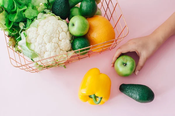 Basket with green fruits and vegetables on pastel pink background