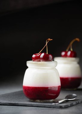 Kavanozda böğürtlensiz tatlı. Siyah tahtada kirazlı İtalyan panna cotta, koyu beton arka plan..