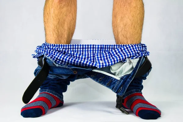 Man has his pants and underwear down  at the ankles.Man in front of white background has pants pulled down.