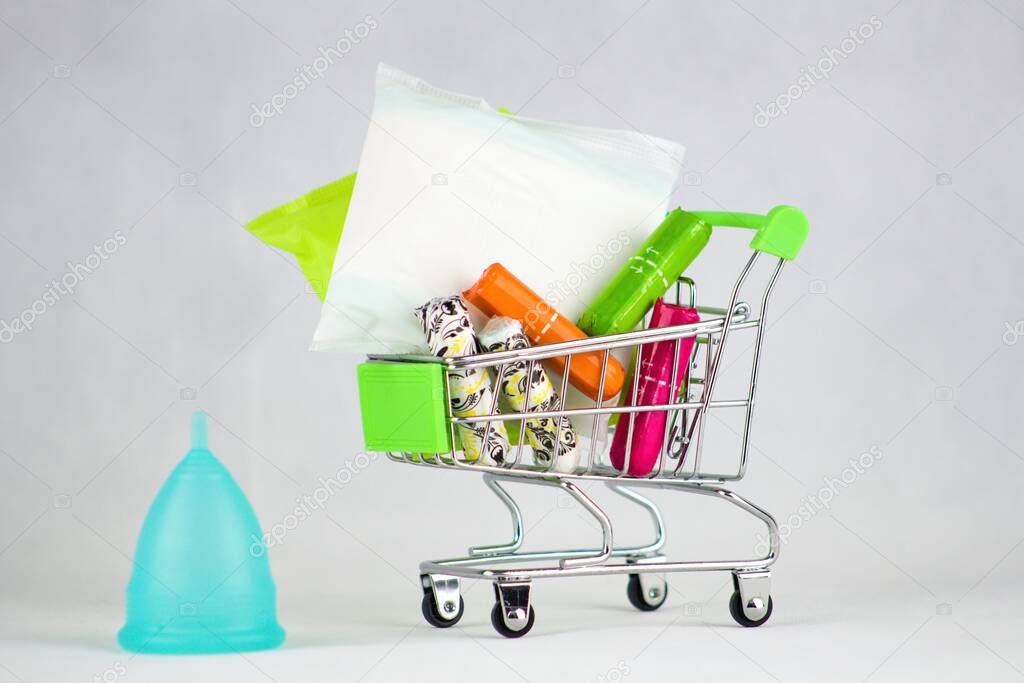 Blue menstrual cup and sanitary napkins and tampons in shopping cart.Menstrual cup - a modern aid during menstruation for a modern woman versus  sanitary napkins and tampons.