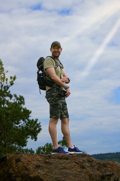 A man - a hiker - stands on top of a mountain.A hiker with outdoor equipment stands on top of a mountain and looks around the surrounding countryside in summer.