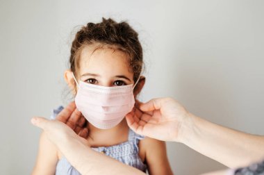 Coronavirus maskeli kız dışarı çıkmaya hazırlanıyor. Annenin elleri bulaşıcılığı önlemek için kızına maske takıyor.