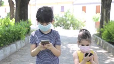 Çocuklar parkta bir bankta oturup telefonla oynuyorlar. Bulaşıcı hastalıklara karşı koruyucu maskeli çocuklar teknolojiye takıntılılar.