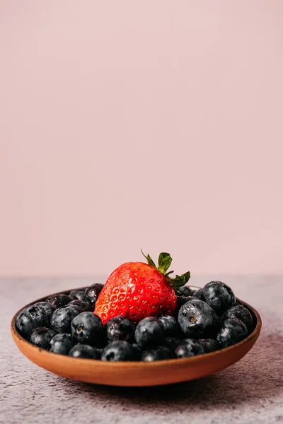 Rustic handmade plate full of blueberries with a strawberry on top ready to eat placed on a cement textured background and pink background — Stock Photo, Image