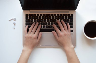 Beyaz bir masaüstüne yerleştirilen ve kahve fincanı ve kablosuz kulaklıklarla çevrili bir bilgisayar dizüstü bilgisayarı üzerinde yazı yazan yaratıcı kadın elinin kesilmiş bir görüntüsü. Düzenli, minimalist bir çalışma alanı.