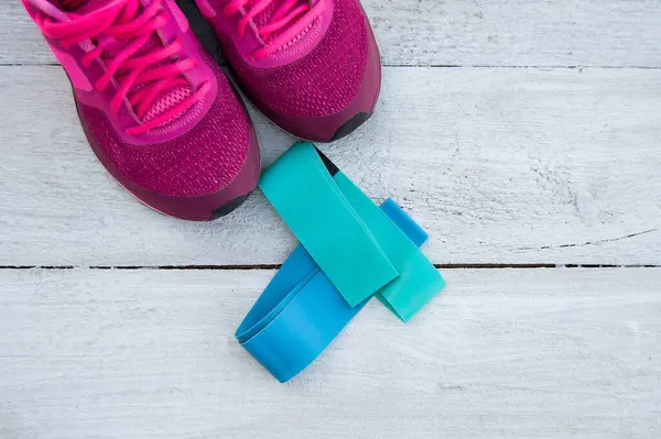 Blue and turquoise elastic rubber bands for fitness and bright pink sneakers on a white wooden background. Flat lay. The view from the top. Ideal for training at home on your own