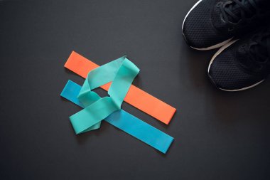 Formda kalmak için çok renkli lastik bantlar ve siyah arka planda siyah spor ayakkabılar. Farklı renklerde lastik bantlar. Kırmızı, mavi ve turkuaz. Evde eğitim için ideal..