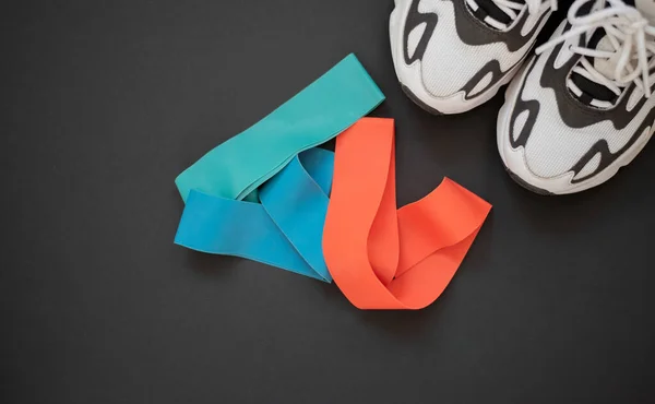 Multi-colored elastic rubber bands for fitness and white and black sports shoes on a black background. A set of rubber bands of different colors - red, blue and turquoise. Ideal for training at home.