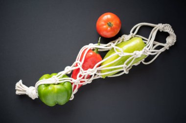 Siyah arka planda parlak taze sebzelerle örülmüş ip torbası. Yeşil, sarı ve kırmızı biberli örgü bir çanta. Çevre dostu çanta. Üst görünüm.