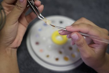Diş teknisyeni cımbız kullanarak dişlere seramik malzeme uygular ve diş macunu arkaplanını fırçalar. Diş teknolojisi yakın plan.
