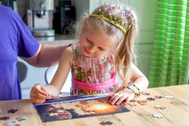 Bir kız masada oturur ve evde bir yapboz toplar. Kameraya bakar ve güler.