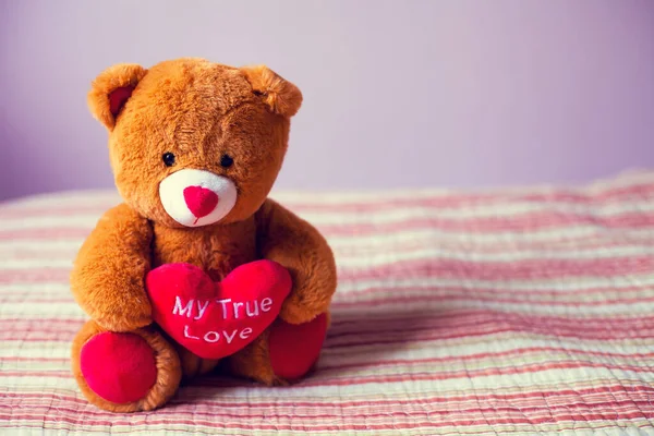 Pençelerinde kalp olan bir oyuncak ayı, duvarın arka planında bir yatakta oturur.