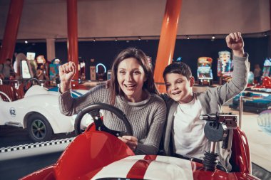 Happy smiling mother and son sitting on toy car. Spending holiday together with family. Entertainment center, mall, amusement park. Family rest, leisure concept. clipart