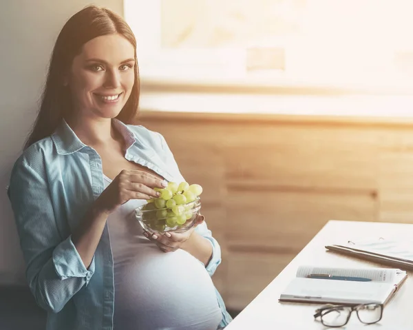 Sonriente joven embarazada comiendo uva en un tazón . — Foto de Stock