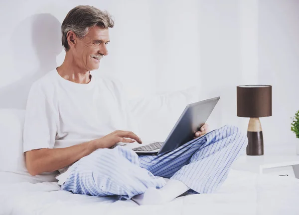 Alter Mann benutzt Laptop während er im Bett sitzt. — Stockfoto