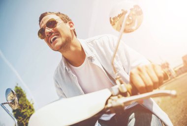 Cadde boyunca Scooter sürme gülümseyen genç adam. Güneş gözlüklü genç adam. Yaz seyahat. Rahat moda. Açık hava etkinliği. Mutlu yaz aylarında seyahat etmek. Sokakta sürme. Motosiklet ile adam.