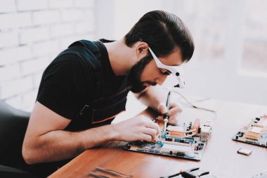Genç adam Pc. tamircisi anakart tamiri sakallı. Araçları ile çalışan. Bilgisayar donanım. Büyüteç Camı. Havya. Dijital aygıt. Laptop üstünde okul sırası. Elektronik cihazlar kavramı.