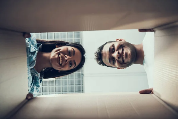 Smiling Young Couple looking Inside Caton Box. Bottom view of Beautiful Couple looking inside Cardboard Box. Young Couple Enjoy Unpacking Cardboard Boxes in New Apartment. Relocating Concept