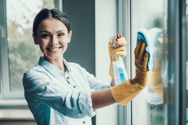 Young Smiling Woman Washing Window with Sponge. Happy Beautiful Girl wearing Protective Gloves Cleaning Window by spraying Cleaning Products and wiping with Sponge. Woman Cleaning House