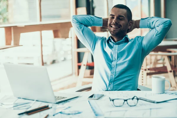 Gülümseyen Genç İşadamı Ofiste Dinleniyor. Dizüstü bilgisayar ofiste. İş adamı yapılan işten memnun kaldı. Masada Elleri Başının Arkasında Oturarak Rahatlayan Adam. Kurumsal Yaşam Tarzı Konsepti