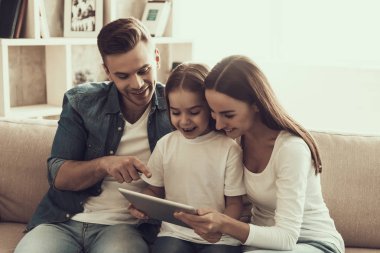 Genç ve mutlu bir aile tablet kullanarak koltukta oturuyor. Güzel Kadın ve Yakışıklı Adam Koltukta Oturuyor ve Sevimli Küçük Kız 'a Tablet Gösteriyor. Ebeveynler ve çocuk birlikte. Aile ve Ebeveynlik Konsepti