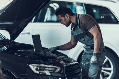 Otomatik tamir dükkanında çalışan araba tamircisi. Dizüstü bilgisayar kullanarak işi araba önünde duran gri üniformalı yakışıklı ciddi beyaz tamirci portresi. Onarım hizmet anlayışı.